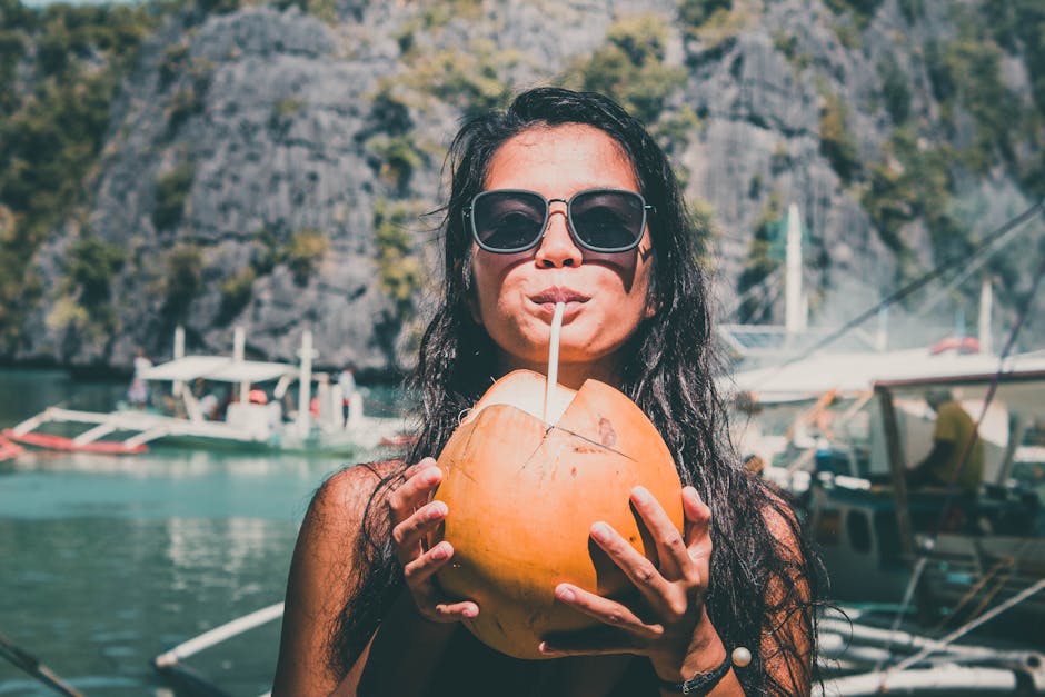 A woman in sunglasses enjoying a fresh coconut drink by the water, with boats and mountains in the background